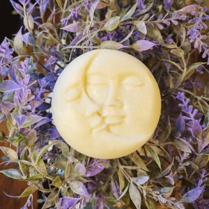 May include: A white soap bar with a sculpted design of two faces, one with closed eyes and the other with an open mouth, resembling the sun and moon. The soap bar is resting on a bed of purple lavender flowers.