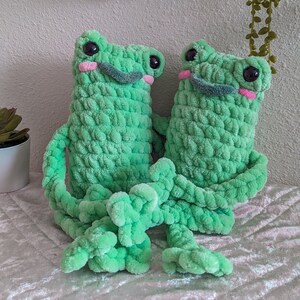 May include: Two crocheted green frog plushies with pink cheeks and black eyes. The frogs are sitting together with their arms around each other.