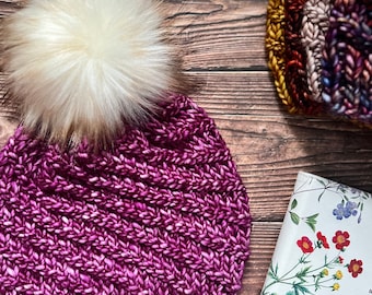 May include: A purple knitted beanie with a white faux fur pom pom. The beanie has a small brown leather tag with the word "Ctenog"