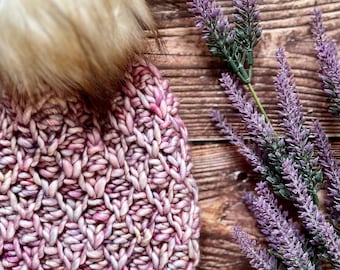 May include: A pink and purple knitted beanie with a fluffy white pom pom. The beanie is sitting on a wooden surface with a sprig of lavender.