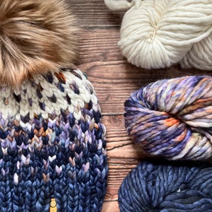 May include: A blue, white, and brown knitted hat with a fluffy brown pom pom. The hat is made with chunky yarn and has a label that says "Standout". There are four skeins of yarn in the background, one white, one blue, one purple and one multi-colored.