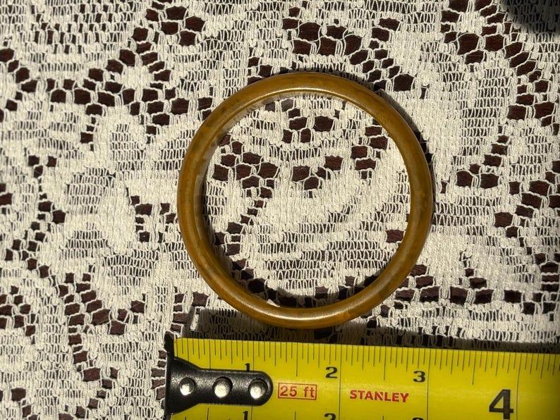 K&ouml;nnte beinhalten: Ein runder, hellbrauner Armreif. Der Armreif besteht aus einem festen Material und hat eine glatte, polierte Oberfl&auml;che. Der Armreif hat einen Durchmesser von ca. 7,6 cm. Der Hintergrund ist ein wei&szlig;es Spitzendesign.