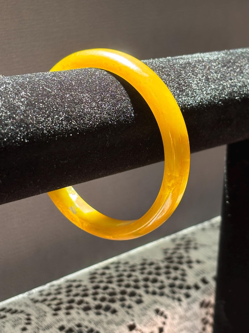 K&ouml;nnte beinhalten: Ein durchscheinendes, gelb-oranges Armreif-Armband. Das kreisf&ouml;rmige Armband besteht aus einem festen Material und hat eine glatte, polierte Oberfl&auml;che. Das Armband wird auf einer schwarzen, strukturierten Oberfl&auml;che pr&auml;sentiert.