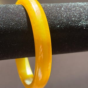 K&ouml;nnte beinhalten: Ein durchscheinendes gelbes Armreif-Armband. Das Armband ist ein massives Band aus poliertem Material mit einem warmen, goldenen Farbton. Das Licht scheint durch das Armband und erzeugt einen Glanzeffekt. Das Armband wird vor einem dunklen, strukturierten Hintergrund pr&auml;sentiert.