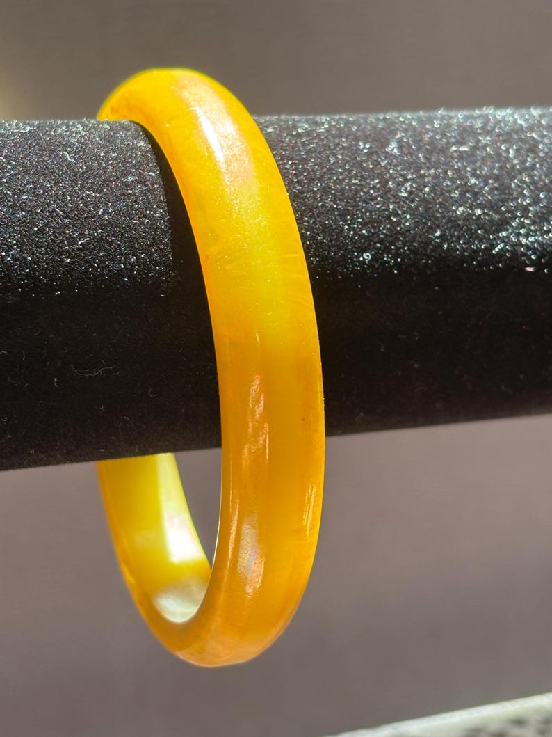 K&ouml;nnte beinhalten: Ein durchscheinendes gelbes Armreif-Armband. Das Armband ist ein massives Band aus poliertem Material mit einem warmen, goldenen Farbton. Das Licht scheint durch das Armband und erzeugt einen Glanzeffekt. Das Armband wird vor einem dunklen, strukturierten Hintergrund pr&auml;sentiert.