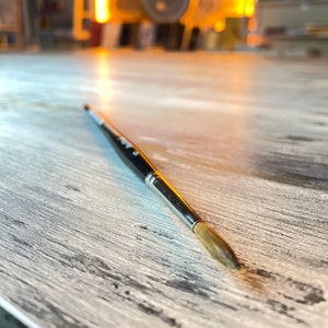 Puede incluir: Primer plano de un pincel negro y plateado con una punta de cerdas marrones. El pincel está sobre una superficie de madera clara.