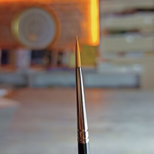 May include: A close-up of a paintbrush with a thin, pointed, yellow bristle tip and a silver metal handle.