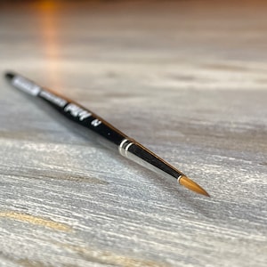 Może przedstawiać: Zbliżenie na czarny i srebrny pędzel z okrągłym końcem. Pędzel leży na jasnoszarym drewnianym podłożu.