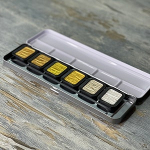 Könnte beinhalten: Ein Metall-Aquarellfarben-Set mit sechs Näpfchen mit metallischem Gold- und Silberfarbton. Die Näpfchen sind in einer Reihe in einem silbernen Etui mit einem weißen Deckel angeordnet.