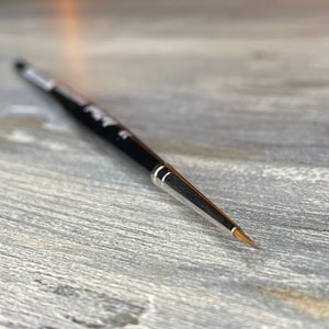 Puede incluir: Primer plano de un pincel negro y plateado con una punta fina. El pincel está sobre una superficie de madera gris claro.