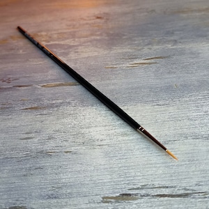 May include: A black paintbrush with a long, thin handle and a fine, golden-colored tip. The brush is lying on a light gray wooden surface.