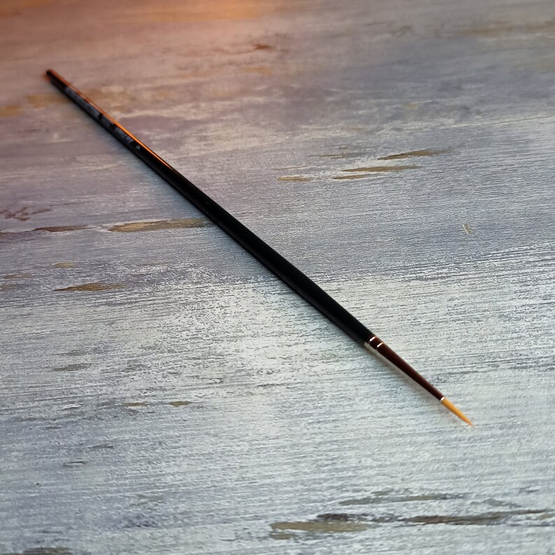 May include: A black paintbrush with a long, thin handle and a fine, golden-colored tip. The brush is lying on a light gray wooden surface.