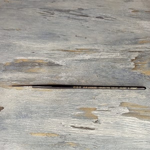 Puede incluir: Un pincel de artista de punta fina con mango negro y letras doradas. El pincel está etiquetado como "000 Grumbacher - Maestro Kolinsky 10 - Japan Made". El pincel está diseñado para pintar con detalle.