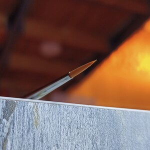 May include: A close-up of a fine-tipped paintbrush with a silver handle and a golden brown bristle tip.