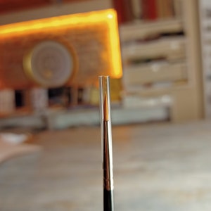 May include: A close-up of a paintbrush with a thin, golden-brown bristle and a clear plastic handle. The brush is standing upright on a table.