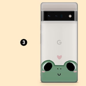 May include: A white phone case with a green frog illustration and a pink heart. The frog is looking up with its mouth open and has large black eyes. The phone case has a black camera lens and the Google logo.