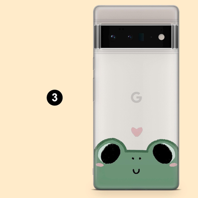 May include: A white phone case with a green frog illustration and a pink heart. The frog is looking up with its mouth open and has large black eyes. The phone case has a black camera lens and the Google logo.