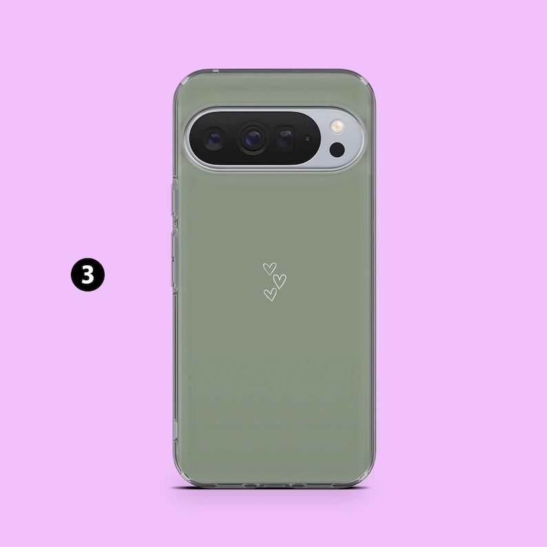 May include: A sage green phone case with a clear border. The case features a camera cutout and a small design of three white hearts. The phone case is shown against a pink background.