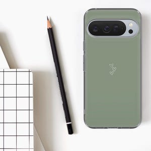 May include: A sage green phone case with a clear border, featuring a camera cutout and three small white hearts. A black pencil and a white notebook with a black grid pattern are also visible.