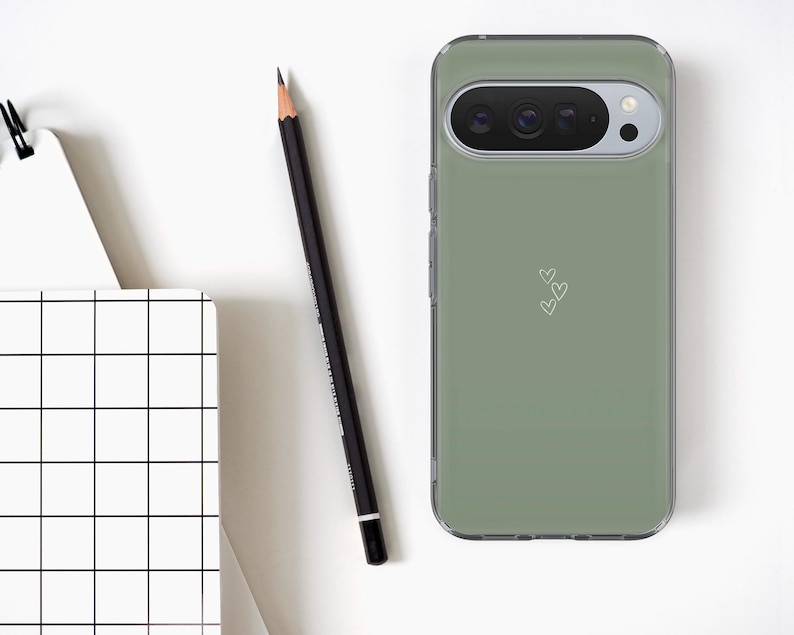 May include: A sage green phone case with a clear border, featuring a camera cutout and three small white hearts. A black pencil and a white notebook with a black grid pattern are also visible.
