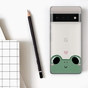 May include: A white phone case with a green frog illustration and a pink heart. The frog has black eyes and a smile.