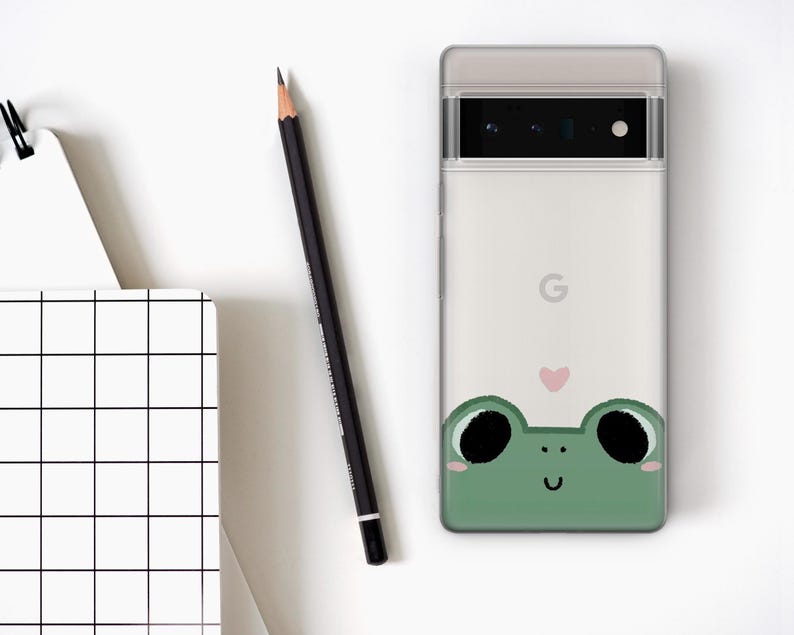 May include: A white phone case with a green frog illustration and a pink heart. The frog has black eyes and a smile.