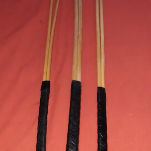 May include: Three wooden polo mallets with black leather grips. The mallets are made of light-colored wood and have a dark, textured grip wrapped around the lower portion. The background is a solid red color.
