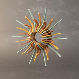 May include: A multi-coloured, abstract sunburst wall decor. The design features a central circular opening surrounded by layered wooden elements in shades of brown, yellow, and green. The radiating elements extend outward, creating a dynamic, three-dimensional effect.