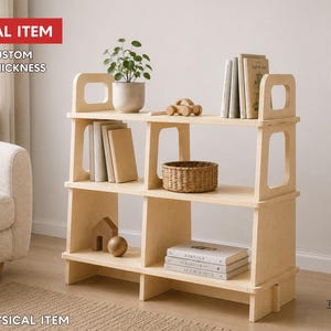 May include: A light-colored wooden bookshelf with three tiers and cut-out handles on the top side panels. The shelves hold books, a potted plant, a small wooden car, a woven basket, a wooden house shape, and a wooden sphere. Text reads DIGITAL ITEM.