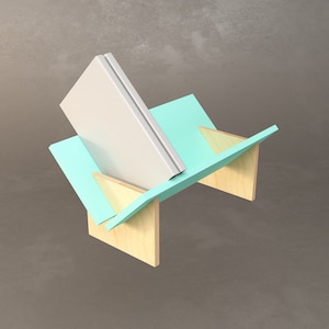 May include: A white book is displayed on a wooden book stand with a teal blue base. The stand is angled and the book is propped up at an angle.