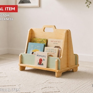 May include: A light-colored wooden book display stand featuring a central handle, a low front panel in a soft sage green, and multiple slots holding various children's books. The text DIGITAL ITEM, FREE CUSTOM SIZE & THICKNESS, and NO PHYSICAL ITEM is visible.