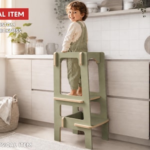 May include: A sage green wooden learning tower with a step stool design, featuring light wood accents on the edges. The structure includes a platform for standing and a lower step, providing a stable elevated surface for reaching kitchen counters.