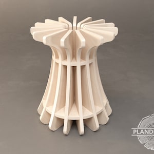 Op de afbeelding: Een houten kruk met een uniek, geometrisch ontwerp. De kruk is gemaakt van meerdere stukken hout die aan elkaar zijn bevestigd om een ​​sterburst-patroon te creëren. De kruk is lichtbruin van kleur en heeft een gladde afwerking.