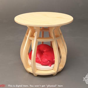 May include: A light wood side table with a round top and a unique, open-sided base. Inside the base is a red sculpture resting on a white cushion. The table is approximately 20 inches tall.