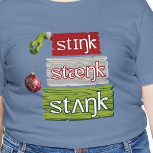 May include: A light blue t-shirt with the words "stink, staenk, stank" in a stacked design. The design includes a green hand holding a Christmas ornament. The shirt is made of a soft material.