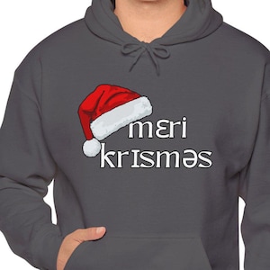 May include: A dark gray hoodie with a red and white Santa hat graphic and the text "mεri krisməs". The hoodie has a front pocket and a drawstring hood. The design is festive and suitable for the holiday season.