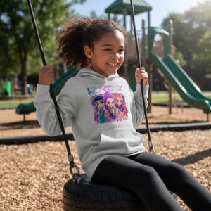 Peut inclure: Un sweat à capuche gris clair avec le mot "HUNTRIX" en lettres bleues et violettes. En dessous, un graphisme de trois personnages de dessins animés. Le sweat à capuche a une poche avant et est porté par une personne sur une balançoire.