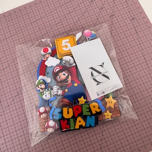 Op de afbeelding: Een kleurrijke Super Mario-thema taarttopper met de naam "KIAN" en het nummer "5". Het ontwerp bevat Mario, Luigi, paddenstoelen, sterren en een bakstenen achtergrond. De topper zit in een doorzichtige plastic zak.