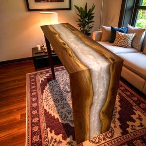 Puede incluir: Una consola de madera artesanal con un diseño de río de resina blanca. La mesa presenta un acabado de vetas de madera natural y mide aproximadamente 152 cm de largo. La mesa tiene un diseño moderno y es adecuada para una sala de estar o entrada.