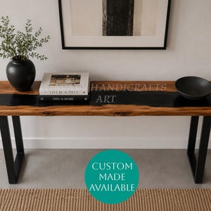 Puede incluir: Una mesa consola rectangular con una superficie de madera natural y una incrustación central de resina negra, sostenida por dos patas de metal negro en forma de U. Un jarrón negro con vegetación, dos libros y un cuenco negro están encima.