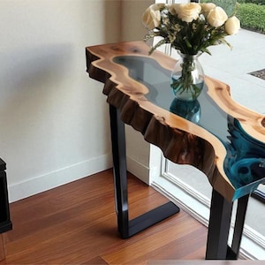 Puede incluir: Una consola de borde vivo con un río de epoxi transparente y patas de metal negro. La mesa tiene un acabado de vetas de madera natural y un jarrón con rosas blancas. La mesa mide aproximadamente 122 cm de largo.