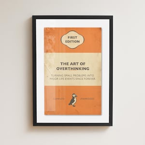 May include: A framed print featuring an orange and cream book cover design with the text "FIRST EDITION," "THE ART OF OVERTHINKING," "TURNING SMALL PROBLEMS INTO MAJOR LIFE EVENTS SINCE FOREVER," "COMPLETE," and "UNABRIDGED," with a small puffin illustration.