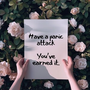 May include: A white rectangular sign with the text "Have a panic attack. You've earned it." is held in front of a backdrop of roses. The sign is held by two hands, with the text in a black, handwritten font.