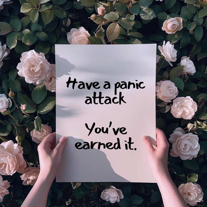 May include: A white rectangular sign with the text "Have a panic attack. You've earned it." is held in front of a backdrop of roses. The sign is held by two hands, with the text in a black, handwritten font.