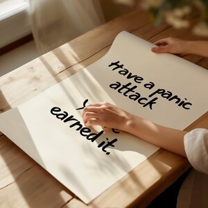 May include: Two off-white sheets of paper on a wooden table with the handwritten text "Have a panic attack" and "You earned it." A person's hands are holding the paper. The lighting is soft and natural.