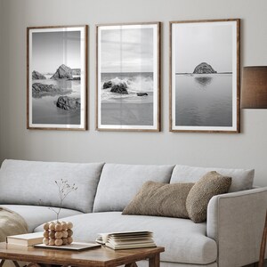 May include: A gray sectional sofa with a throw blanket and a wooden coffee table. Three black and white framed photographs of seascapes hang on the wall above the sofa.