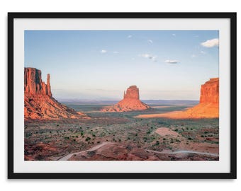 May include: A panoramic view of Monument Valley in Arizona, featuring three red rock formations against a blue sky with white clouds. The landscape is a mix of red rock, sand, and sparse vegetation.