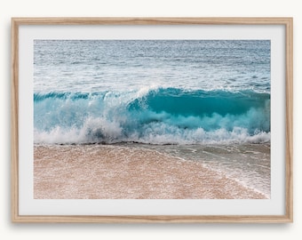 May include: A framed print of a turquoise ocean wave breaking on a sandy beach. The wave is captured mid-break, revealing its translucent interior. The water transitions from deep blue to white foam, contrasting with the tan sand and the light blue of the sea.