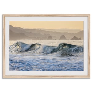 May include: A photograph of a seascape with waves crashing on the shore. The waves are a light blue and green color, and the water is a deep blue. In the background, there are mountains in the distance. The sky is a light yellow color.