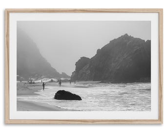 May include: Black and white framed photograph of a beach scene. The image features a sandy beach, ocean waves, and large rock formations. Several people are visible on the beach and in the water. The photograph is enclosed in a light brown wooden frame.